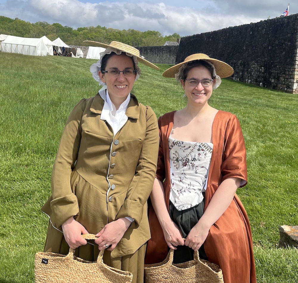 Two people in 1780s women’s clothing stand in front of Fort Frederick. The one on the left wears an olive-drab riding habit. The one on the right wears a russet sack-back gown. /images/2023/annalee_maco.jpg