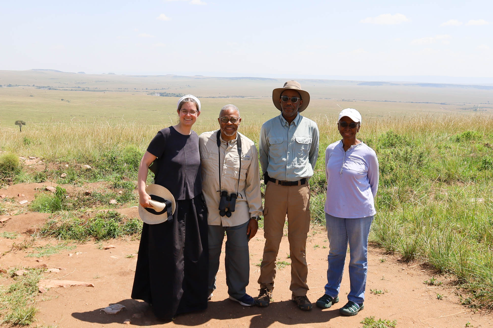 4 people stand on a hill with the savannah laid out behind us. Me dressed in a dark grey t-shirt and black skirt, holding a hat, a man with short grey hair in a kakhi shirt and grey pants with binoculars, a taller man in a hat with a blue shirt and brown pants, and a woman in a white baseball cap, lavender shirt, and jeans. /images/2023/baltimore_friends_on_safari.jpg