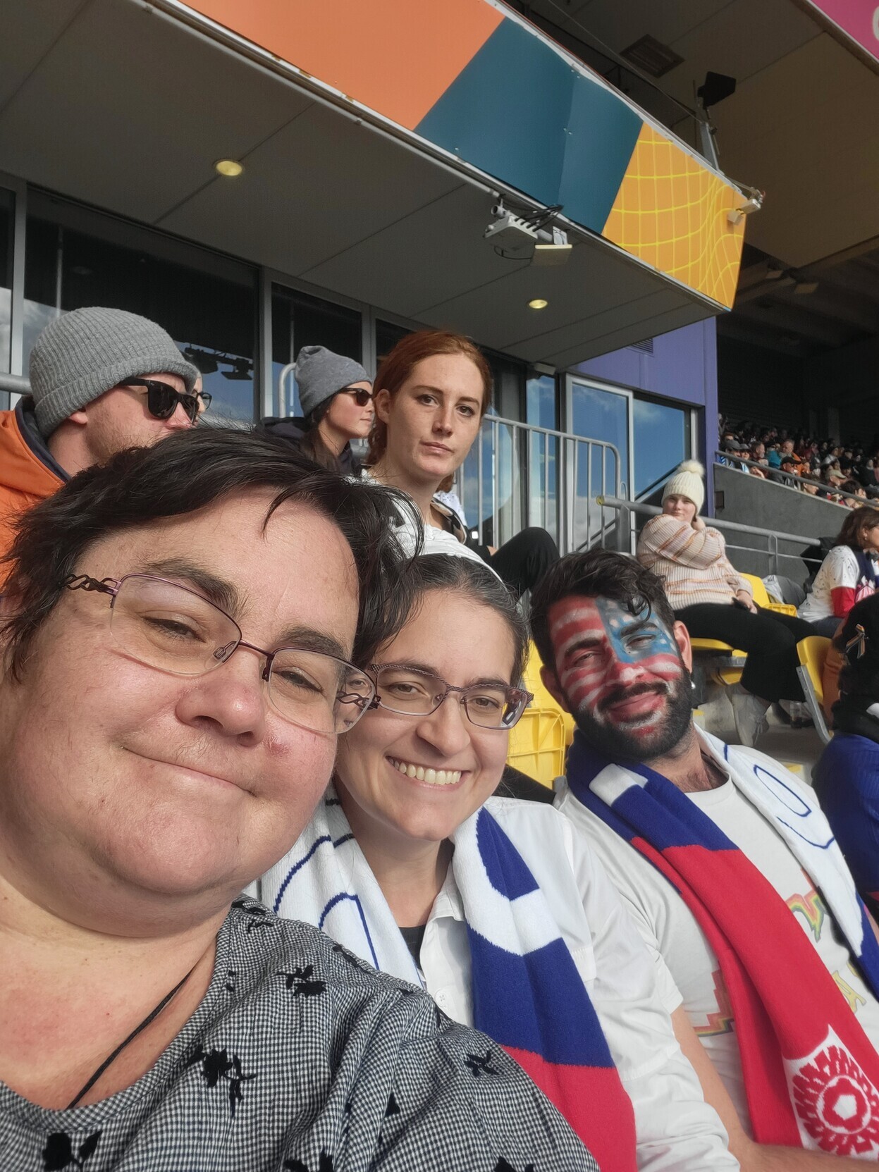 3 people sit in the stands at Sky Stadium in Wellington, New Zealand, taking a selfie. Nearest is Brenda, with short brown hair and a checked shirt. Next is me, in a white shirt with a USA scarf draped over my neck. Finally, my brother, a bearded man with wavy hair who has painted his face into an American flag and also wears a USA scarf. /images/2023/brenda_maco_ross.jpg