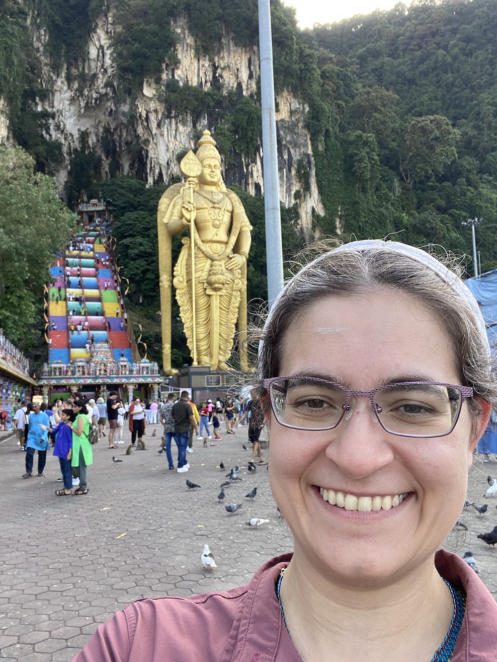 I smile in front of a cliff face that shows many caves. There are colorful stairs leading much higher than I want to climb. There’s also a tall golden statue of the Hindu god Marugan and to the side a temple is visible. /images/2023/maco_batu_caves.jpg