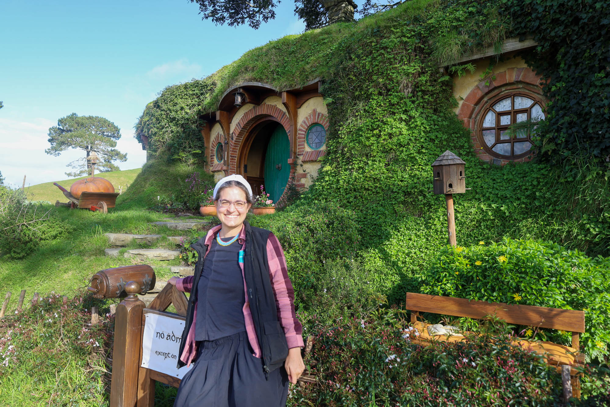 I stand in front of Bag End, its green door visible behind, and the “no admittance except on party business” sign at the gate, though I block part of the sign. I’m layered up, with a black vest over pink and maroon plaid flannel shirt over grey t-shirt, with a black skirt. /images/2023/maco_hobbiton.jpg