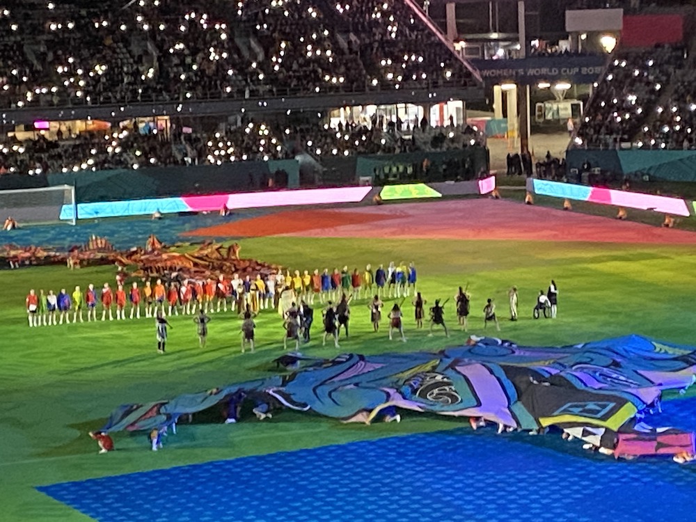 Evening view of Eden Park with the field covered to protect it. A haka is being performed in front of representatives from all the teams. The crowd uses their cell phone lights the way people used to use lighters in olden days. /images/2023/world_cup_opening_ceremony.jpg